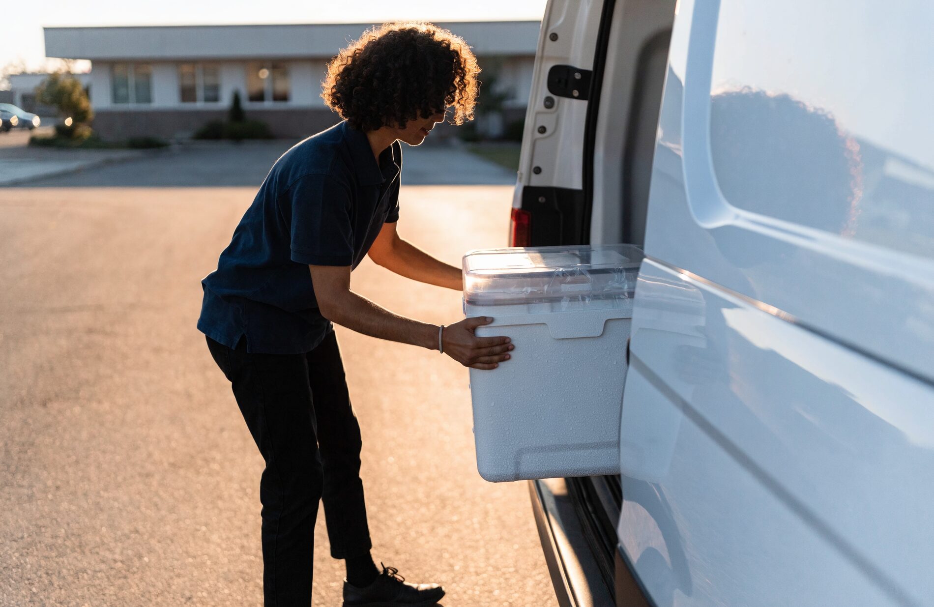 A medical courier loads an insulated specimen cooler into a white courier van outside a clinic at first light